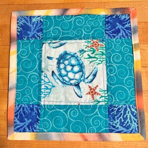 May include: A square quilted fabric mat featuring a central panel with a blue sea turtle, orange starfish, and green seaweed. The border consists of teal fabric with white swirling patterns and dark blue corner squares with coral motifs.