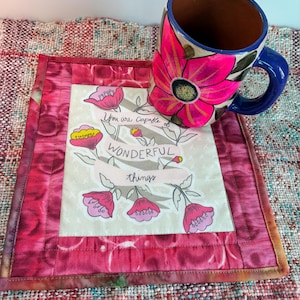 May include: A handmade mug with a large pink flower design and a blue handle rests on a quilted coaster. The coaster features a floral design with the text "You are capable of wonderful things."