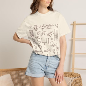 May include: A cream-coloured short-sleeve t-shirt featuring dark brown line art illustrations of football-themed icons, including a trophy, helmet, goal post, and megaphone, with the text "washington football" printed in the centre.