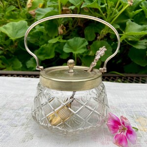 以下が含まれることがあります： 銀色の金属製のハンドルと蓋が付いた、透明なカットガラスの砂糖入れ。小さなスプーンが蓋の上に置かれています。ボウルはダイヤモンド模様です。ピンクの花がボウルの隣にあります。