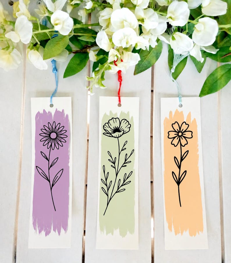 May include: Three rectangular paper bookmarks featuring black line art of flowers centered over vertical brushstrokes of color. The left bookmark has a purple stroke, the center a light green, and the right a peach stroke, each with a colored tassel.