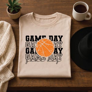 Pu&ograve; includere: T-shirt beige con la scritta "GAME DAY" impilata verticalmente, con una grafica di pallone da basket arancione. Le parole sono in grassetto nero. L'ultima riga di testo recita "game day" in corsivo.