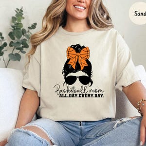 Pu&ograve; includere: T-shirt color sabbia con grafica di una donna con occhiali da sole e fiocco a forma di pallone da basket. Il testo recita "Basketball mom ALL.DAY.EVERY.DAY."