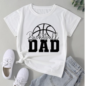 Pu&ograve; includere: T-shirt bianca con grafica di pallone da basket nero e la scritta "Basketball DAD". La maglietta &egrave; abbinata a scarpe da ginnastica bianche e jeans blu. La maglietta &egrave; annodata in vita.