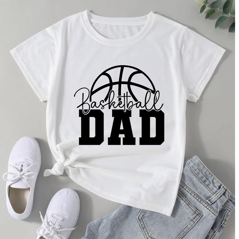 Pu&ograve; includere: T-shirt bianca con grafica di pallone da basket nero e la scritta "Basketball DAD". La maglietta &egrave; abbinata a scarpe da ginnastica bianche e jeans blu. La maglietta &egrave; annodata in vita.