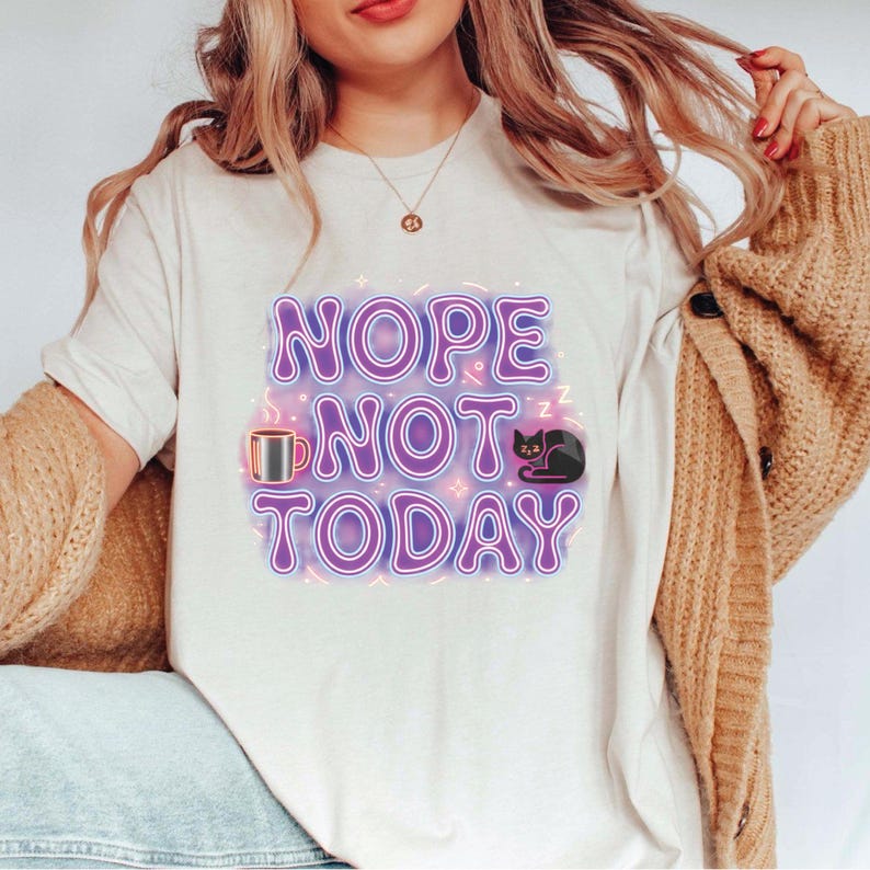 K&ouml;nnte beinhalten: Cremefarbenes T-Shirt mit dem Schriftzug "NOPE NOT TODAY" in lilafarbenen Buchstaben. Das Design zeigt eine Kaffeetasse und eine schlafende schwarze Katze mit Neon-Effekt.
