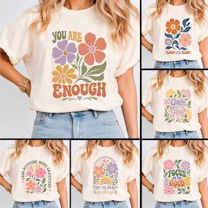 Pode incluir: Uma t-shirt de manga curta cor de creme com seis designs gr&aacute;ficos diferentes. Cada design apresenta ilustra&ccedil;&otilde;es florais coloridas combinadas com textos inspiradores como You Are Enough, Bloom Baby Bloom, Grow With The Flow, Less Attitude More Gratitude, Find The Beauty In Everyday e Focus On The Good.