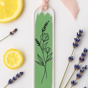 May include: A clear acrylic bookmark featuring a vertical sage green brushstroke design with a black line art illustration of a wildflower stem. The bookmark is topped with a soft pink fabric ribbon tied through a circular hole at the top.