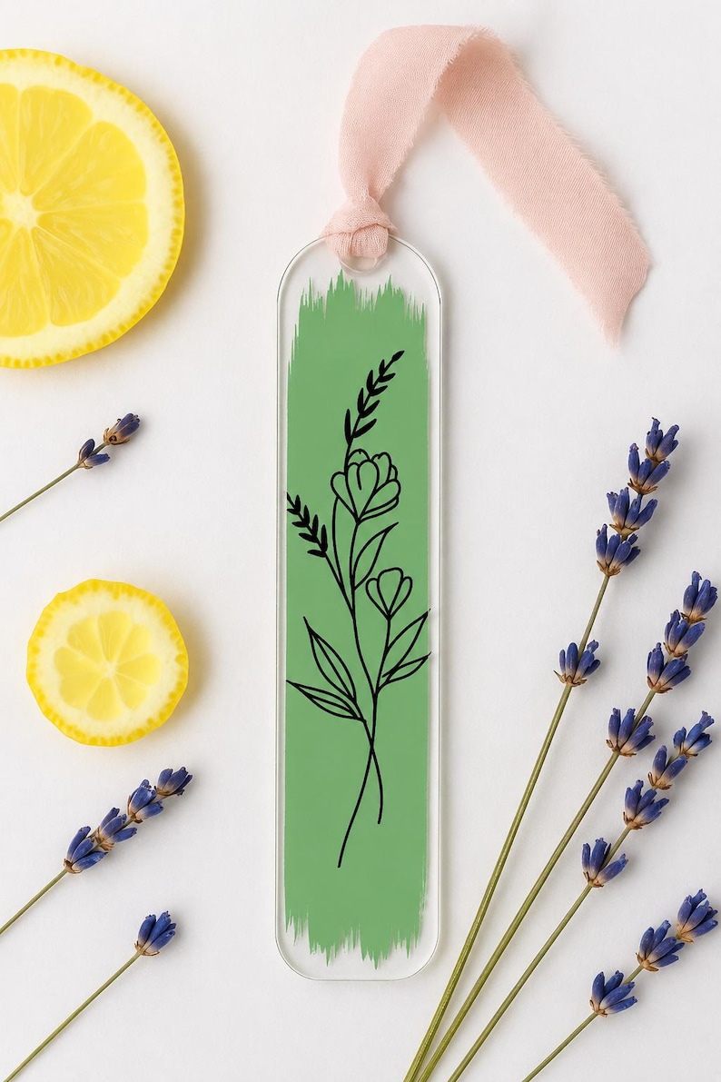 May include: A clear acrylic bookmark featuring a vertical sage green brushstroke design with a black line art illustration of a wildflower stem. The bookmark is topped with a soft pink fabric ribbon tied through a circular hole at the top.