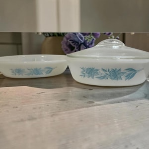 May include: Two white milk glass dishes with a blue floral pattern. One dish is a shallow oval bowl, and the other is a round casserole dish with a clear glass lid. Both pieces feature a repeating design of blue flowers and leaves on the exterior.