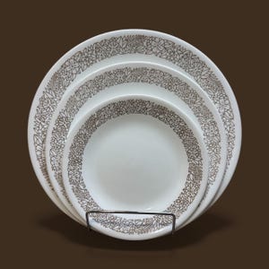 May include: A set of three white ceramic plates with a brown floral and leaf pattern around the rims. The plates are stacked and displayed on a metal stand, showcasing a vintage dinnerware set.