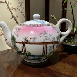 May include: A white ceramic teapot featuring a hand-painted scene of a figure on a horse crossing a bridge. The design includes pink, green, and brown accents with detailed black line work depicting trees and landscape elements, topped with a lid.
