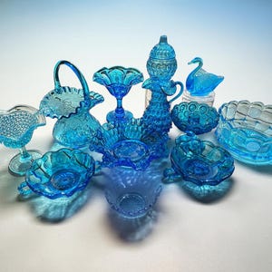 May include: A collection of vintage blue glass decorative items, including bowls, a swan figurine, a basket, and a stemmed dish. The glass pieces have intricate patterns and textures, and are arranged on a light background.