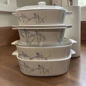 May include: A stack of four white oval ceramic baking dishes with integrated side handles. Each dish features a delicate botanical pattern of purple iris flowers and green stems. Two of the dishes are topped with clear glass lids.