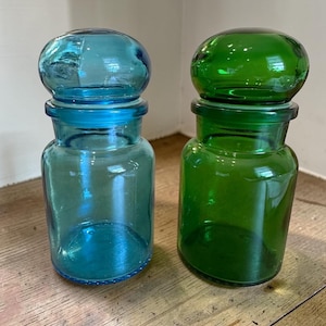 May include: Two glass apothecary jars with rounded lids. One jar is a translucent light blue, and the other is a vibrant emerald green. Both containers feature a cylindrical body with a distinct neck and a matching glass stopper lid.