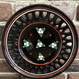 May include: A round black ceramic plate featuring a decorative lattice-style rim with a wavy orange and white border. The center of the plate is adorned with five hand-painted white flowers with green leaves and small orange dots between them.
