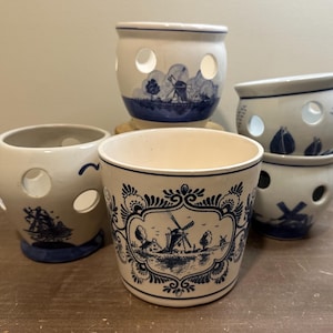 May include: A collection of white ceramic planters with blue windmill and landscape designs. The planters have circular cutouts and vary in size and shape, creating a charming, rustic aesthetic. The designs are reminiscent of traditional Dutch Delftware.