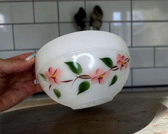 May include: A white milk glass bowl featuring a hand-painted floral pattern of pink flowers with yellow centers and green leaves arranged in a horizontal vine design around the exterior of the rounded vessel.