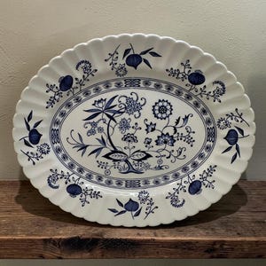 May include: An oval-shaped white ceramic serving platter with a scalloped edge. The platter features a blue floral pattern in the center and a decorative blue border. The design includes stylized flowers, leaves, and fruit motifs in deep blue.