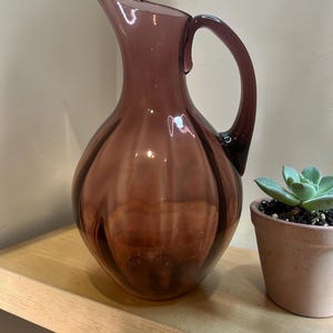 May include: A translucent plum-colored glass pitcher featuring a rounded, bulbous body with vertical ribbed detailing, a slender neck, and a curved handle. The pitcher has a wide, pointed pouring spout and a smooth, polished finish throughout.