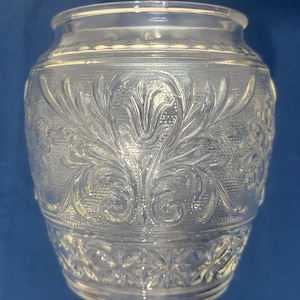 May include: Clear glass jar with embossed floral and geometric patterns. The jar has a wide mouth and a slightly flared rim. The body of the jar features a symmetrical floral design, while the base has a starburst pattern. The jar is likely used for storage or display.