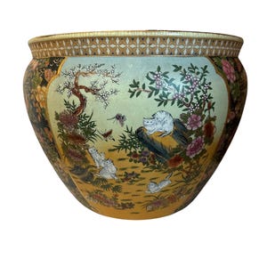 May include: A large, round ceramic planter with a detailed, hand-painted design. The planter features a light green background with a scene of cats, flowers, and trees. The rim has a gold geometric pattern, and the overall color palette includes gold, pink, and black.