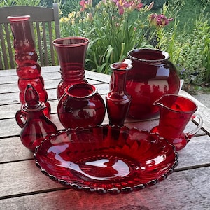 May include: A collection of ruby red glass items including a tall stacked vase, a flared vase, a large round bowl, a small round vase, a slender vase, a small pitcher with a handle, and a wide shallow bowl with a scalloped edge and ribbed base.