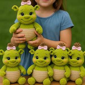 Op de afbeelding: Een jong meisje houdt een groene gehaakte pop vast met een lachend gezicht, een roze strik en een beige luier. Er zijn verschillende vergelijkbare poppen voor haar gerangschikt. De poppen hebben kleine oren en zitten. De achtergrond is vaag groen.