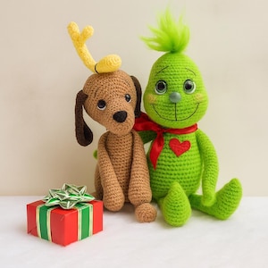 Può includere: Figure all'uncinetto di un cane marrone con corna gialle e una creatura verde con un cuore rosso e una sciarpa rossa. Una scatola regalo rossa con un fiocco verde è di fronte a loro.