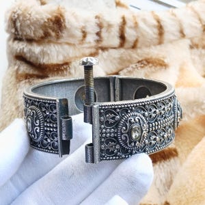 Könnte beinhalten: Silberfarbenes Gelenkarmband mit aufwendigem Filigran und klaren Kristallakzenten. Das Armband verfügt über einen Schraubverschluss und detaillierte Schnörkel. Ein Schmuckstück im Vintage-Stil.