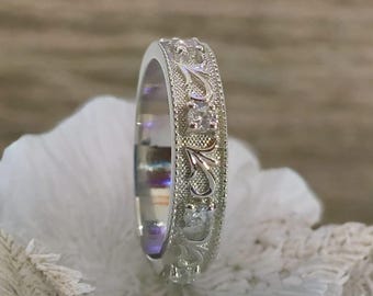 Könnte beinhalten: Ein silberner Ring mit einem aufwendigen floralen Design und eingebetteten klaren Steinen. Das Band des Rings weist detaillierte Gravuren und kleine, funkelnde Akzente auf, die ein zartes und elegantes Aussehen erzeugen. Der Ring ist vor einem weichen, neutralen Hintergrund platziert.