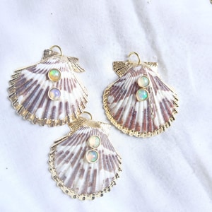 May include: Three seashell pendants with gold-coloured trim and a small loop at the top. Each shell is white and brown with two iridescent opal-like stones. The pendants are displayed on a white surface.