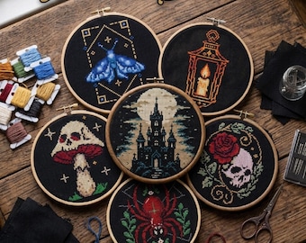 May include: A collection of six cross-stitch embroidery hoops with gothic-themed designs, including a moth, lantern, skull, mushroom, castle, and spider. The hoops are displayed on a wooden surface with embroidery floss and needles.