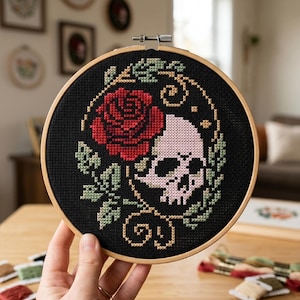 May include: A circular wooden embroidery hoop containing a cross-stitch design on black fabric. The pattern features a large red rose, a pale pink skull, and green leaves, all framed by a thin, golden-brown vine with small decorative swirls.