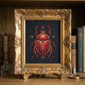 May include: A cross-stitch embroidery of a red beetle centered on a black fabric background, encased in an ornate gold-colored frame. The beetle design features symmetrical red and orange thread work with small cross symbols surrounding the insect.