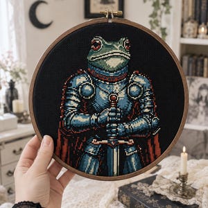 Può includere: Un telaio da ricamo circolare in legno contenente un motivo a punto croce di una rana vestita con un'armatura da cavaliere blu e argento. La rana tiene una spada con entrambe le mani e un mantello rosso sullo sfondo di tessuto nero.
