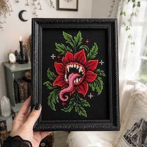 May include: A framed cross-stitch embroidery on black fabric featuring a red carnivorous flower with sharp white teeth and a long pink tongue, surrounded by green leaves and small white and red cross-shaped accents in a black ornate frame.