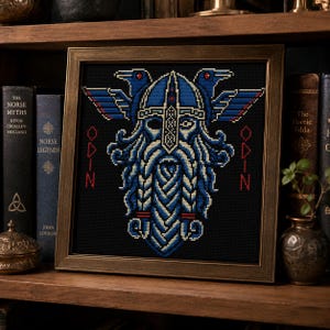 May include: A framed cross-stitch embroidery on a black background featuring a stylized blue and white bearded figure wearing a winged helmet, flanked by two ravens. The name ODIN is stitched in red vertical text on both sides of the central figure.