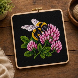 Puede incluir: Bordado de punto de cruz de un abejorro sobre flores de trébol rosa, sobre un fondo negro. La abeja es amarilla y negra, con alas detalladas. El bordado está enmarcado en un aro de madera cuadrado.
