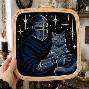 May include: A square wooden embroidery hoop containing a cross-stitch design of a knight in blue armor holding a blue cat. The background is black with gold-colored stitched stars. The knight wears a helmet with a visor and a dark blue cloak.