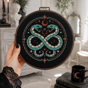 May include: A circular wooden embroidery hoop containing a black fabric piece with a cross-stitch design of two intertwined teal and cream snakes forming an infinity shape, accented by a red crescent moon, small stars, and geometric diamond patterns.