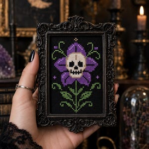 May include: A framed cross-stitch depicting a purple flower with a skull centre, green leaves, and a black background. The frame is ornate and black. The artwork is held in a hand with black nail polish and a silver ring. The theme is gothic and macabre.