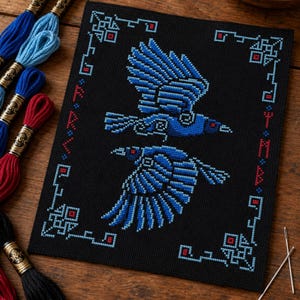 May include: A black cross-stitch fabric piece featuring two blue birds in flight, surrounded by a geometric border in light blue and red. The design includes stylized runic-style characters along the sides, with embroidery floss skeins nearby.