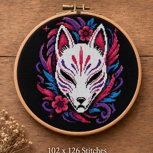 May include: A cross-stitch embroidery of a Kitsune mask in a wooden hoop. The mask is white with red and purple details, surrounded by colorful accents. The text "11 colors" is at the top. The dimensions are 7.3 x 9.0 inches.