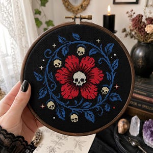 May include: A circular wooden embroidery hoop containing a cross-stitch design on black fabric. The pattern features a large red flower with a central skull, surrounded by a blue leafy vine, small white skulls, and tiny white cross-shaped stars.