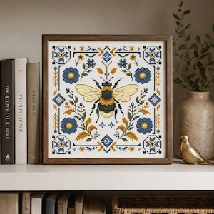 Pode incluir: Um bordado em ponto cruz quadrado numa moldura de madeira escura com um zangão central rodeado por motivos florais e geométricos simétricos em azul e amarelo sobre um fundo de tecido branco, exibido num ambiente doméstico.
