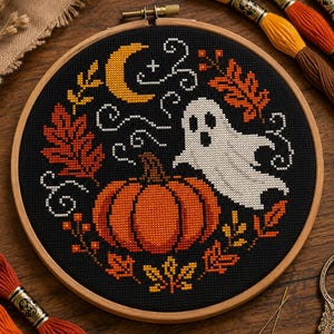 May include: A circular wooden embroidery hoop containing a cross-stitch design on black fabric. The pattern features a central orange pumpkin, a white ghost, a yellow crescent moon, and various orange and yellow autumn leaves with white swirls.