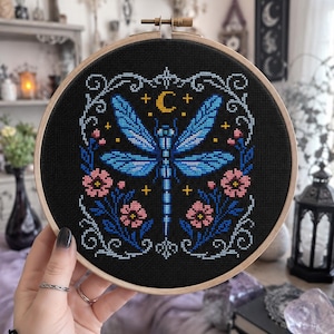 Puede incluir: Un bastidor de bordado circular de madera que contiene un diseño de punto de cruz sobre tela negra. El patrón presenta una libélula azul central, una luna creciente amarilla, pequeñas estrellas y flores rosas con hojas azules, todo enmarcado por un borde de volutas azul claro.