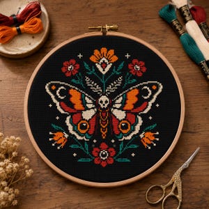 Pode incluir: Um bastidor de bordado em ponto cruz com um design de borboleta colorido, incorporando um motivo de caveira, sobre um fundo preto. A borboleta apresenta tons de laranja, vermelho e branco, complementados por elementos florais. O bastidor de madeira está sobre uma superfície de madeira.