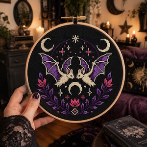 May include: A circular wooden embroidery hoop containing a black fabric piece with a cross-stitch design. The pattern features two cream-colored bats with purple wings, surrounded by crescent moons, stars, and symmetrical purple and pink floral sprigs.